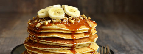 a tall stack of pancakes topped with syrup, chopped nuts, and banana slices