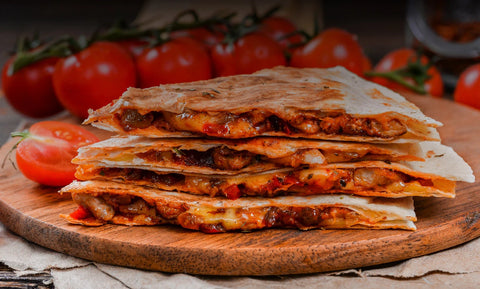 a pile of quesadilla slices sits on a wooden board in front of a row of on-the-fine tomatos