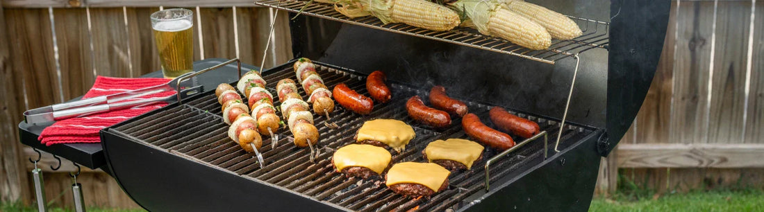 Cheeseburgers, sausages, and vegetable skewers cook on the main grate of a barrel grill. Ears of corn roast on the warming rack. A towel, tongs, and a full drink glass sit on the left side shelf