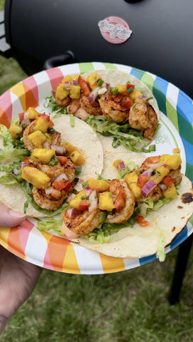 3 open shrimp tacos topped with chopped mangos and onions on a festive paper plate with stripes in a variety of colors