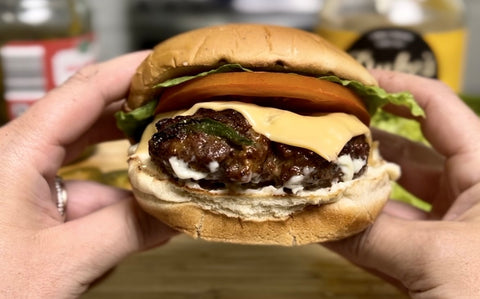 Someone holds a jalapeno chedded stuffed burger in two hands. Melted cheese coming out of the burger. The burger is topped with melted cheese, lettuce, and a thick slice of tomato