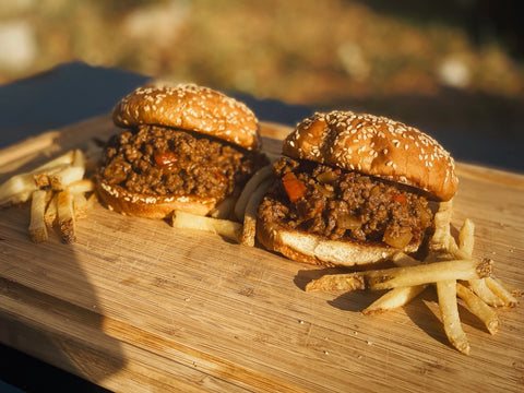 2 stuffed sloppy joes on sesame seed buns sit on a wooden board with french fries scattered around them