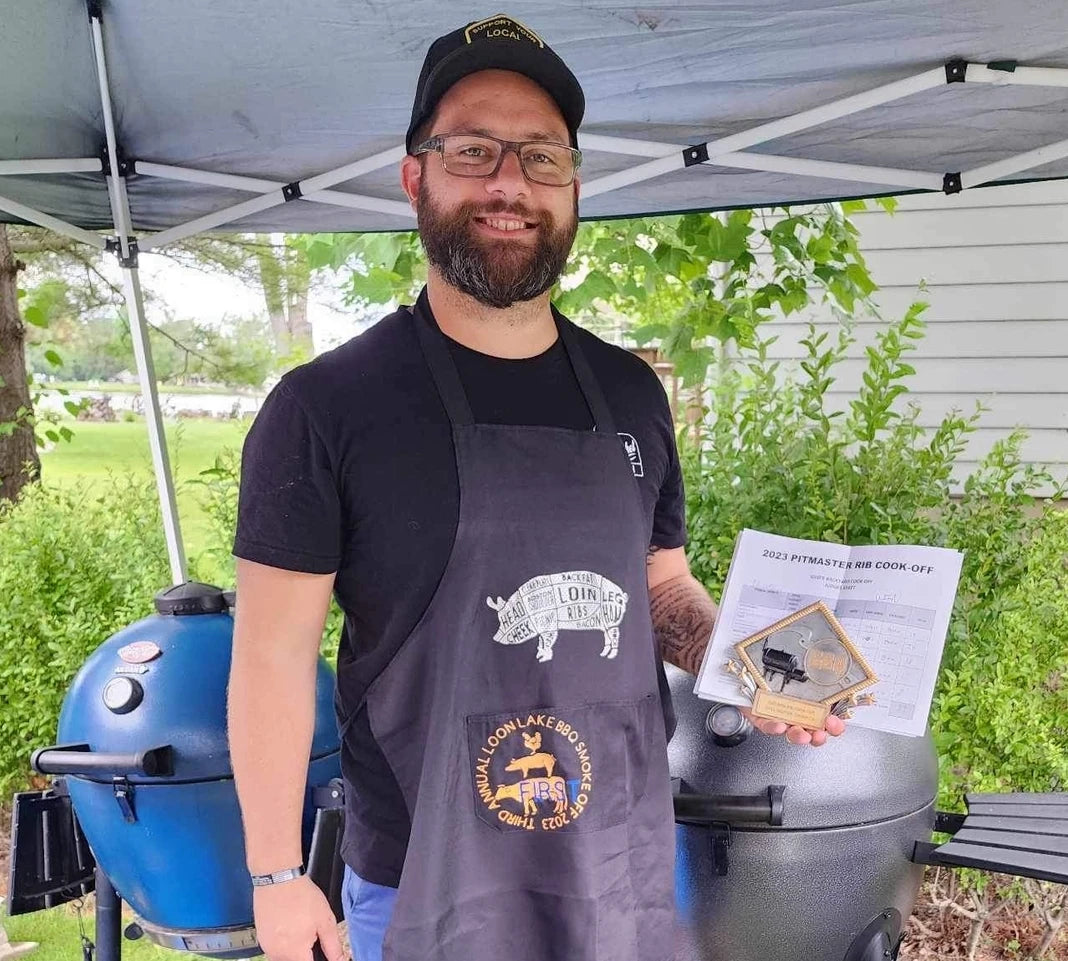 Dalton Rose stands under an outdoor canopy wearing a BBQ competition apron. He's holding up an award he won there.