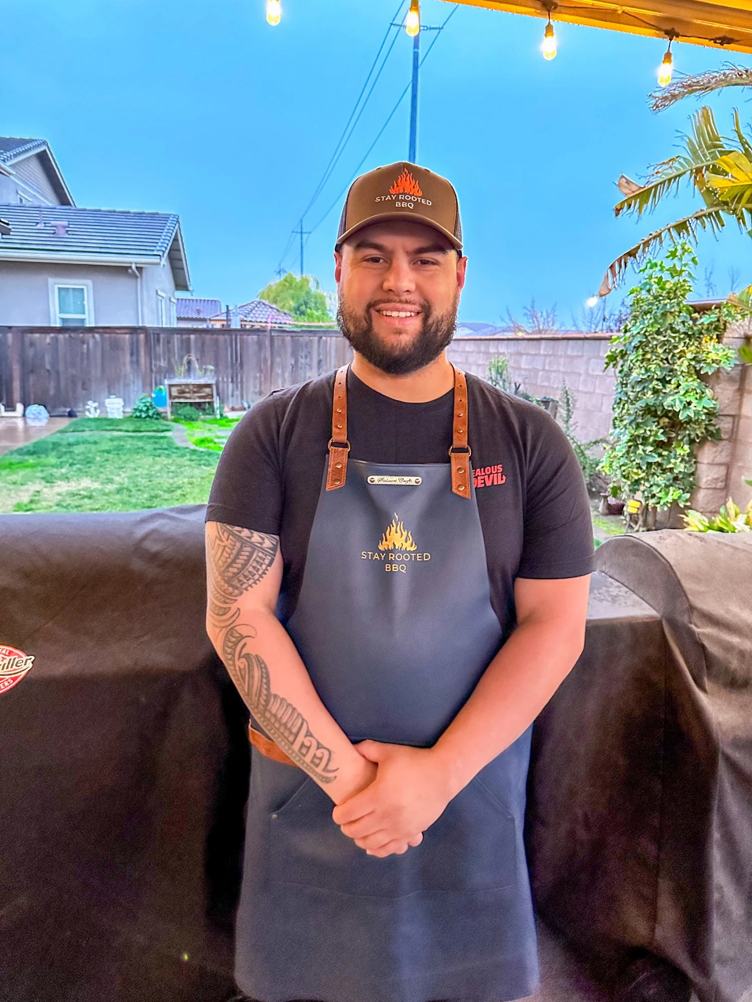 Rob Conrado Jr Stands under an outdoor awning wearing a stayrootedbbq apron