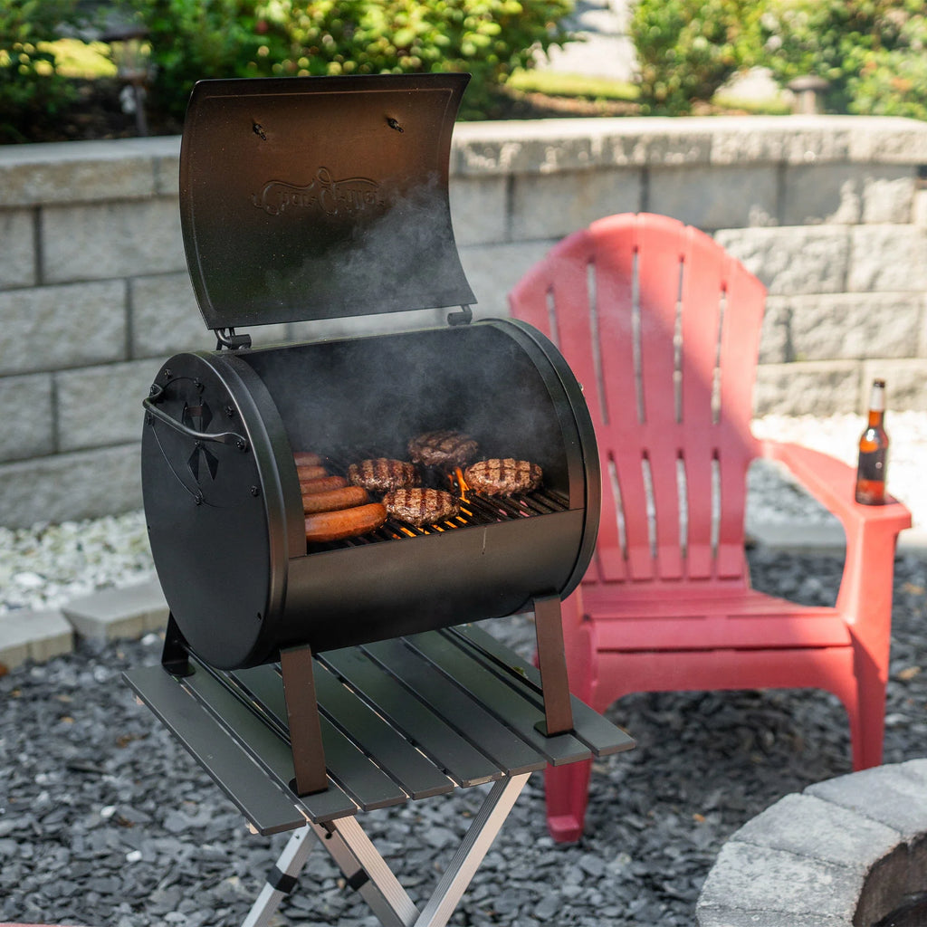 An open grill placed on a folding table outside on a graveled patio in front of a curved retaining wall. A Red chair with a beverage bottle on the arm sits slightly to the right of the grill. 4 burgers and 5 sausages are cooking over flaming coals.