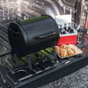 A closed Portable Pro sits on the lowered tailgate of a pickup truck next to a cooler full of ice and bottles  and a tray holding burger buns and cheese slices.