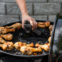 Someone drops blocks of charcoal into an AKORN grill through the access hole in the center of the cooking grate. Cooking chicken legs are scattered around the outer ring of the grate.