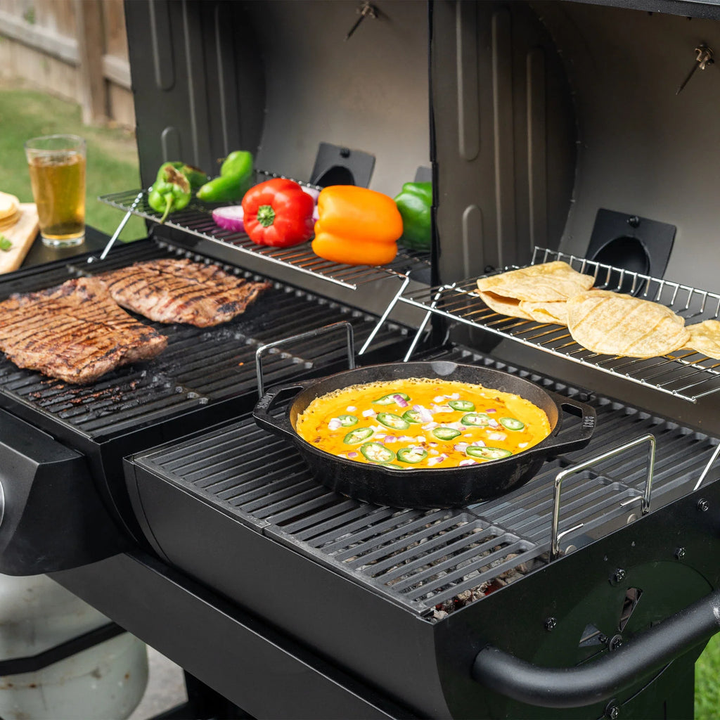 An open Dual-Fuel Pro grill. The gas side (left) has 2 steaks grilling on the main grate and whole bell and chili peppers on the warming rack. The charcoal side (right) has a round cast iron pan of something yellow topped with slices of fresh jalapeno peppers and red onions on the main grate while tortillas warm on the warming rack.