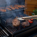 Someone uses long tongs to turn a steak cooking on the charcoal side of the grill. 3 more steaks and cherry tomatoes on the vine are also cooking on the grill. Sweet potato halves cook on the warming rack above them.