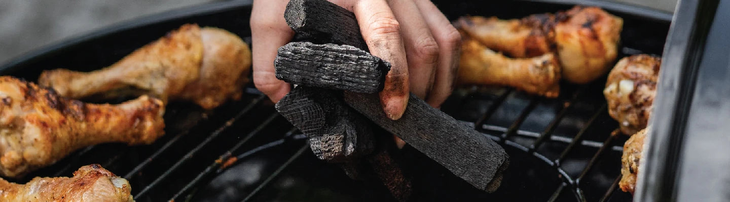 Someone drops unlit Char-Griller lump charcoal into the open center of an AKORN kamado grill. Chicken legs cook on the outer ring of the grill.