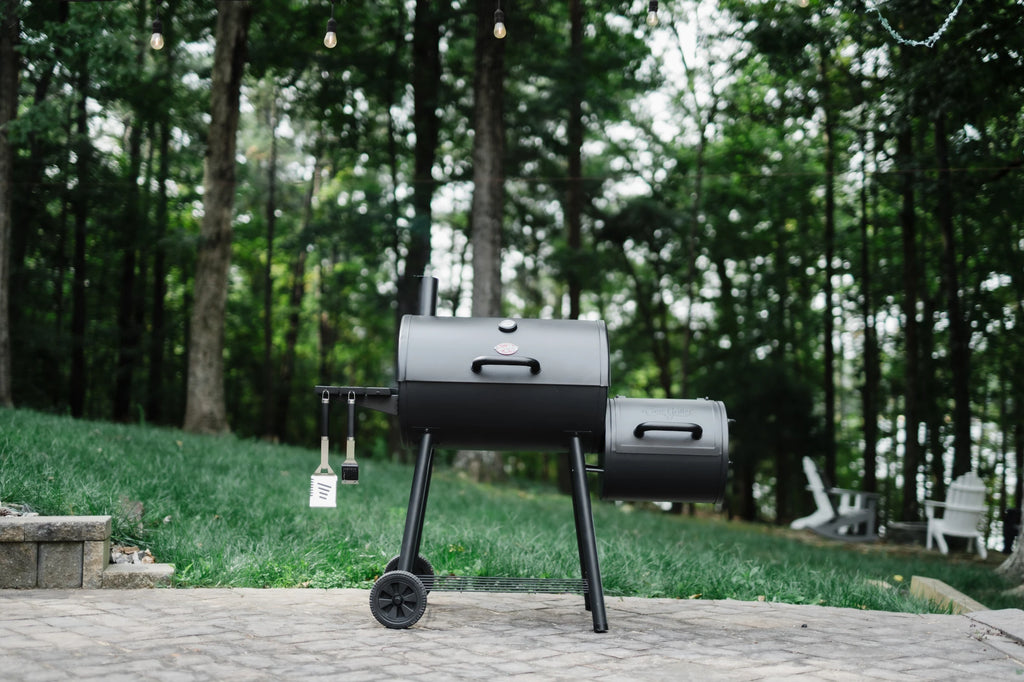 A closed Smokin' Pro grill with attached offset smoker on the right and a side shelf on the left. The smoker is mounted on a cart with 2 large wheels. The grill sits on a paved patio in front of a green lawn and trees.