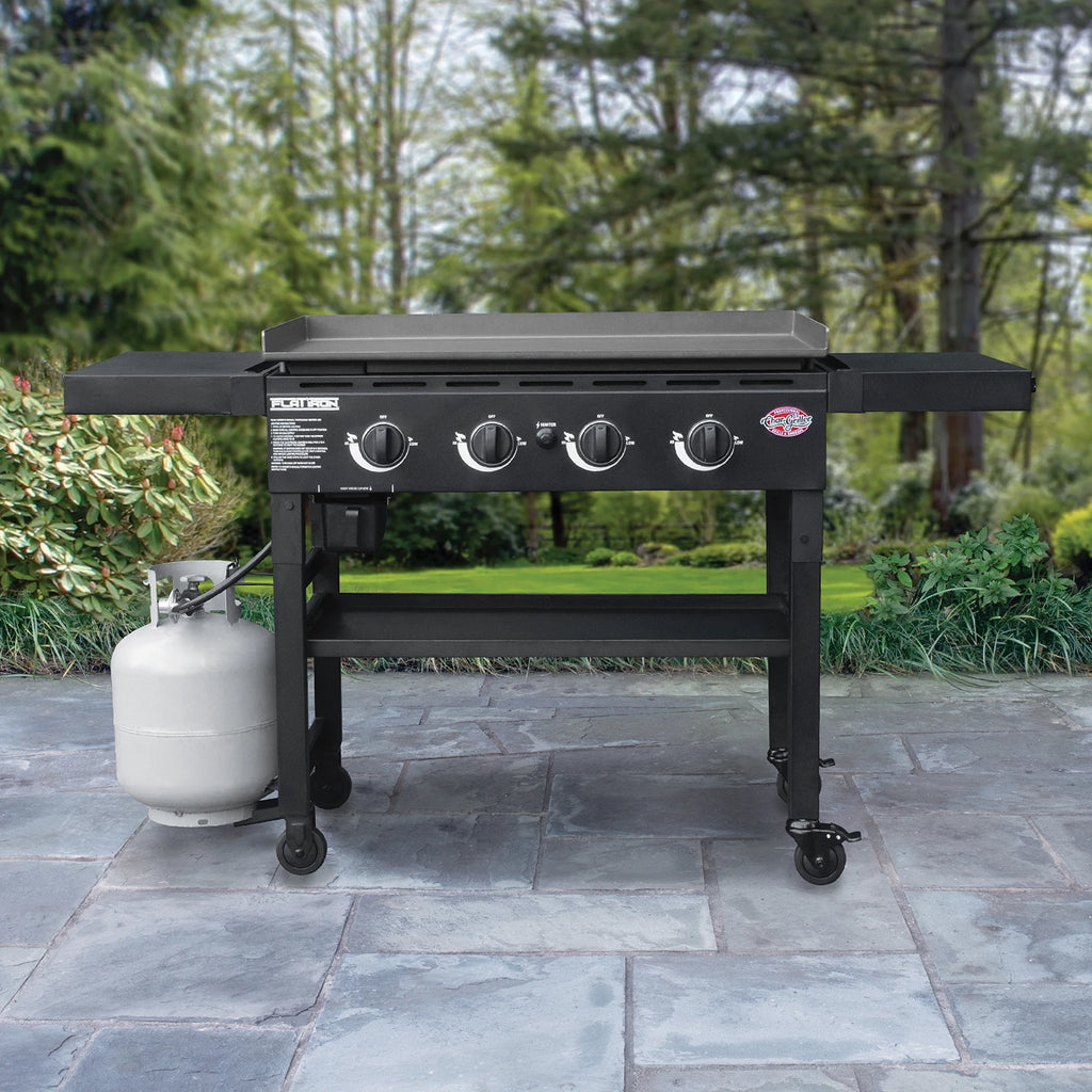 The griddle outside on a paved patio. The griddle is attached to a small propane tank that is sitting on the ground beneath the left side shelf.