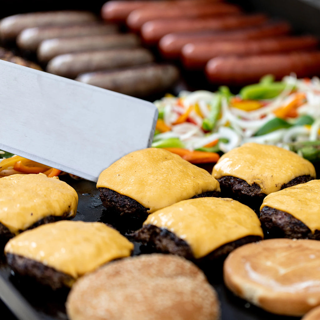 Someone uses a spatula to raise the edge of a cheeseburger patty. Sausages and sliced peppers and onions are visible in the background. Burger buns toast on the griddle in the foreground.