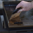 Someone using a grill scraper swipes grease from the griddle into the slide-out grease drawer through a hole in the front-left of the griddle.