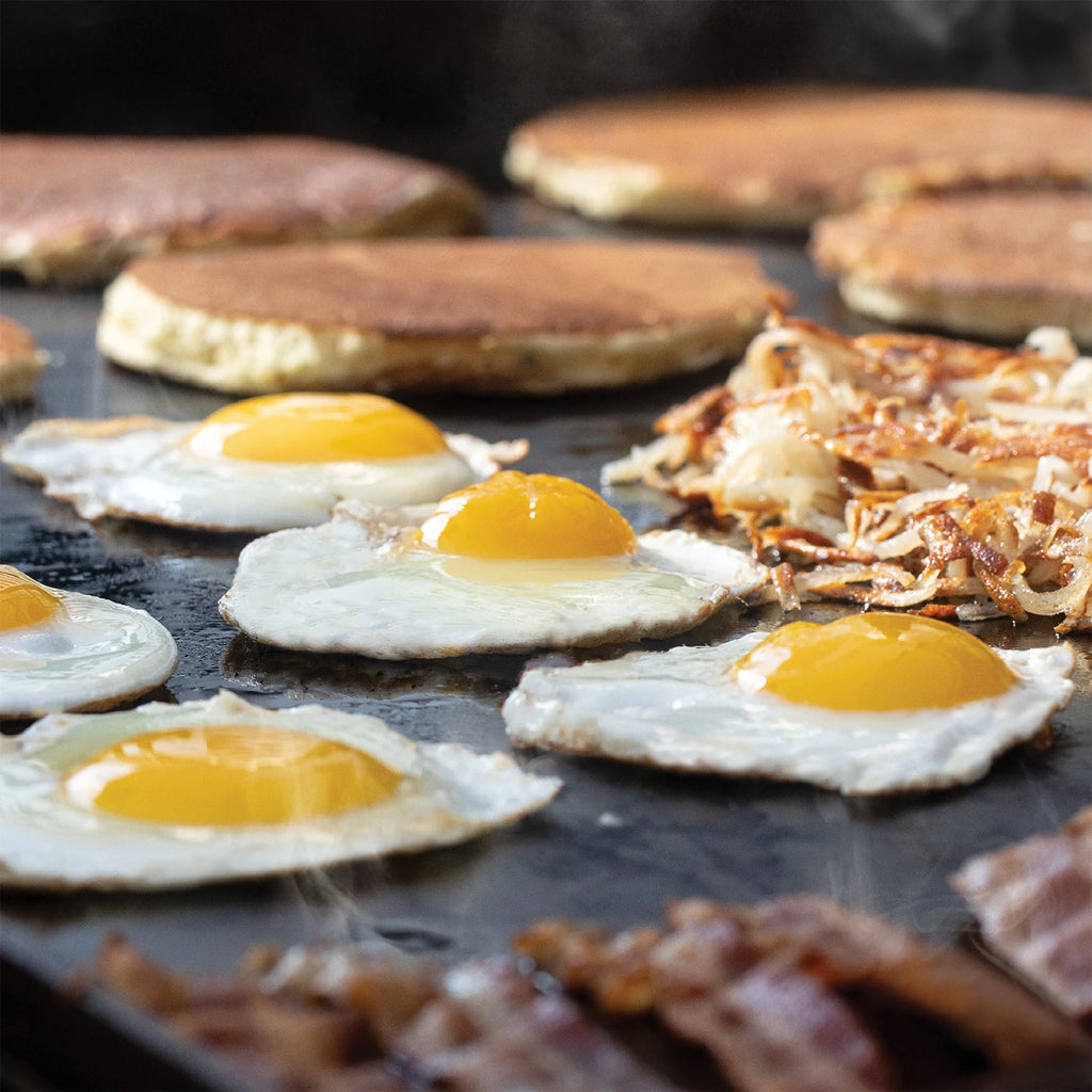 A full breakfast of eggs, pancakes, hash browns and bacon cooking on the griddle.