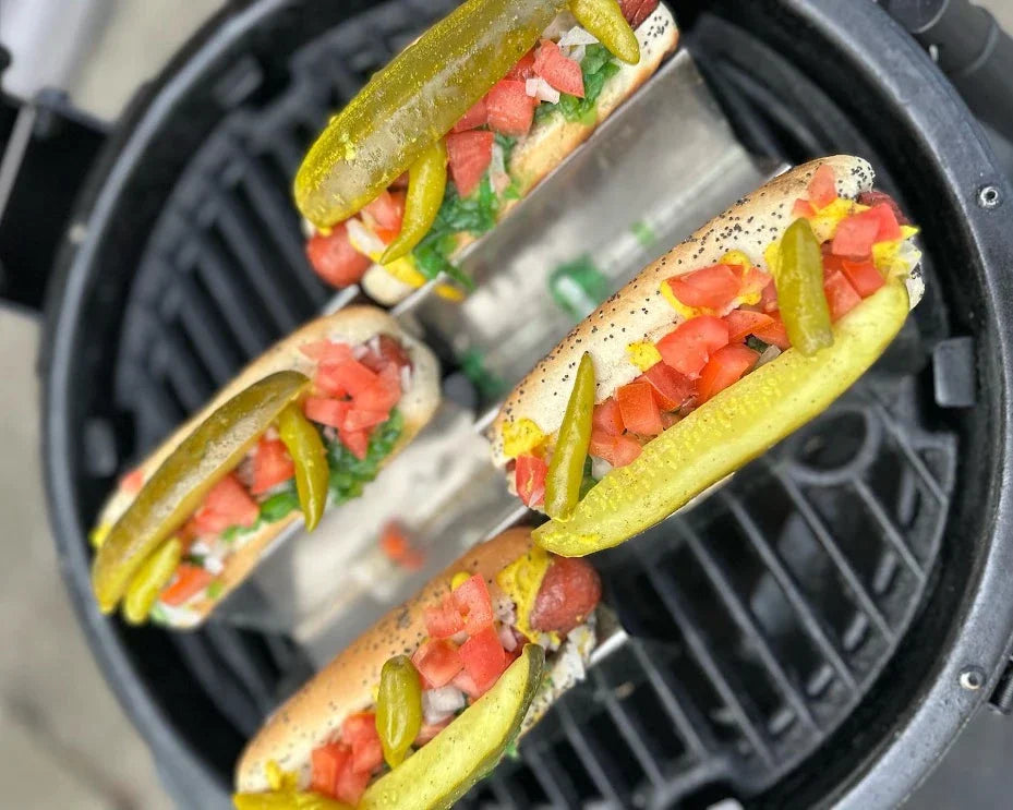 4 hot dogs in seeded buns resting in a metal holder on top of a grill grate. The hot dogs are topped with pickle spears, diced tomatoes and onions, sport peppers, and mustard