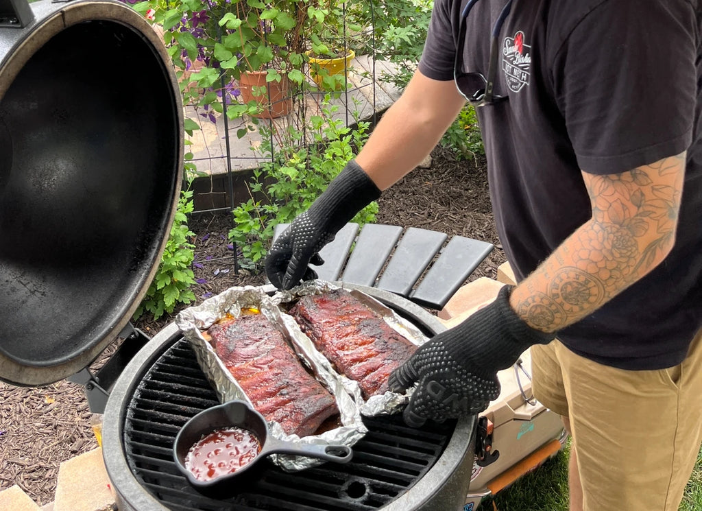 Ambassador Dalton Rose wears grilling gloves as he arranges 2 tinfoil trays of ribs on the grill of a Char-Griller AKORN grill. A cast iron pot of sauce sits on the grill next to the ribs.